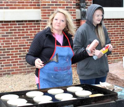 Two women outside a brick building cooking pancakes on a large griddle